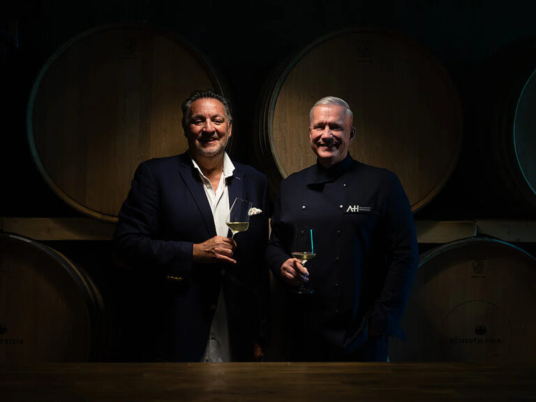 Zwei Männer stehen vor Weinfässern und lächeln in stilvoller Kleidung.