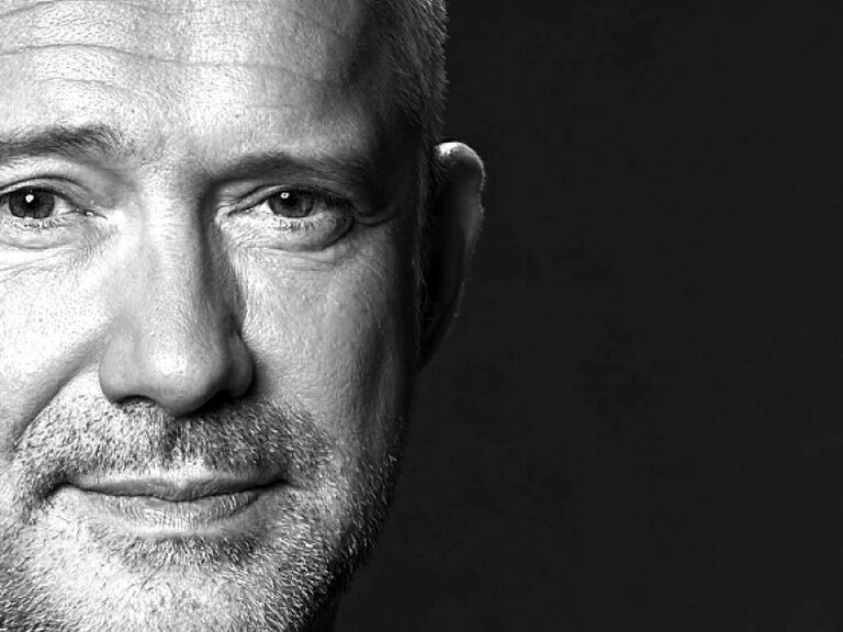 Ein stilvolles Schwarzweiß-Porträt von Alexander Herrmann welches die Struktur seines Anzugs und Gesichts betont.